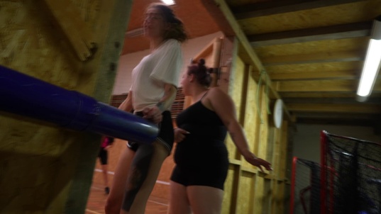 indoor gym,wooden beams,blue padded bar,fluorescent lighting,women standing,athletic wear